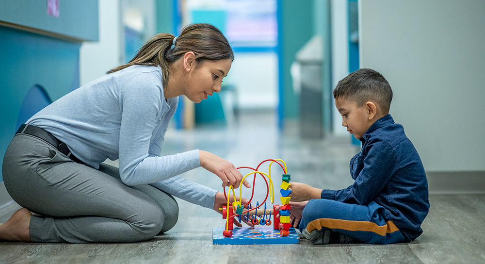 Una joven latina está sentada en el suelo frente a un niño latino. Entre ellos hay un juguete colorido en el que ambos están interesados.