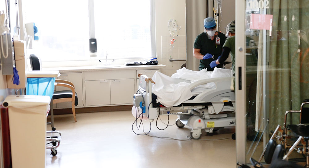 En la habitación de un hospital, un proveedor con bata azul y una mascarilla atiende a un paciente que no se puede ver.