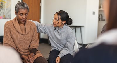 una joven negra consuela a una mujer negra mayor que está sentada en una silla con las manos juntas